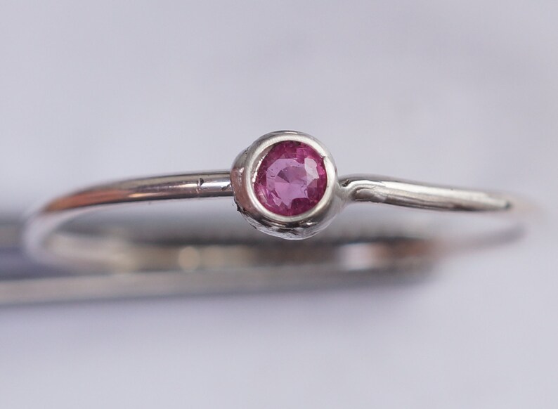 K&ouml;nnte beinhalten: Ein silberner Ring mit einem kleinen, runden, rosafarbenen Edelstein, der in einer Zargenfassung gefasst ist.