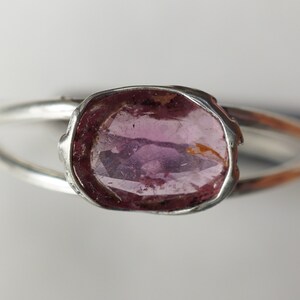 K&ouml;nnte beinhalten: Ein silberner Ring mit einem rosafarbenen Edelstein, der in einer Zargenfassung gefasst ist. Der Ring hat ein schlichtes Band und der Edelstein ist oval.