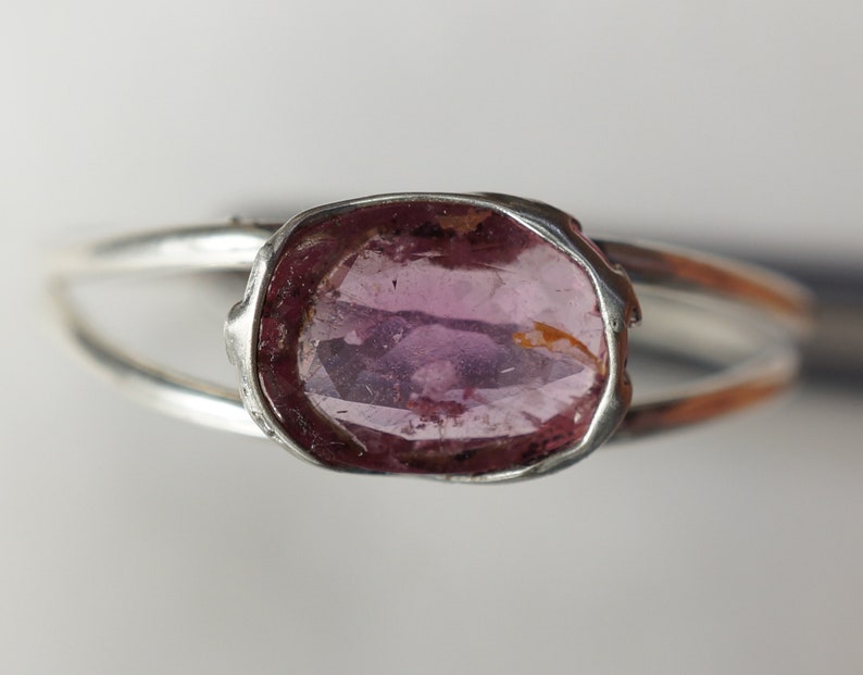 K&ouml;nnte beinhalten: Ein silberner Ring mit einem rosafarbenen Edelstein, der in einer Zargenfassung gefasst ist. Der Ring hat ein schlichtes Band und der Edelstein ist oval.