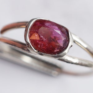 Puede incluir: Un anillo de plata con una piedra ovalada rosa engastada en un engaste de bisel. El anillo tiene una textura martillada.