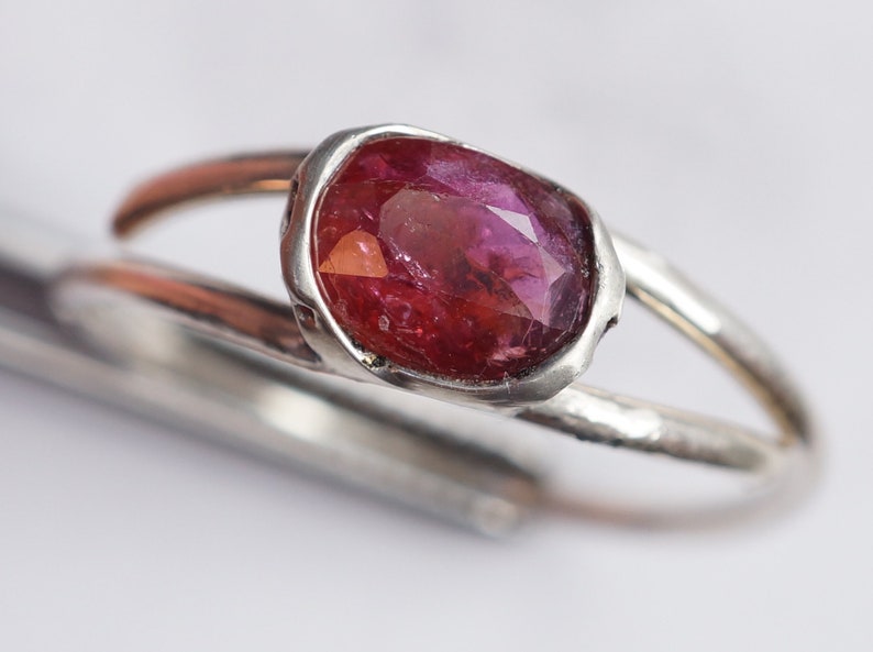 Puede incluir: Un anillo de plata con una piedra ovalada rosa engastada en un engaste de bisel. El anillo tiene una textura martillada.