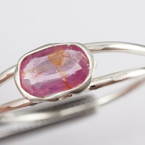 Puede incluir: Un anillo de plata con una piedra preciosa ovalada de color rosa engastada en un engaste de bisel. El anillo tiene una banda doble.