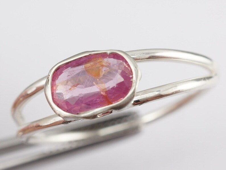 Puede incluir: Un anillo de plata con una piedra preciosa ovalada de color rosa engastada en un engaste de bisel. El anillo tiene una banda doble.