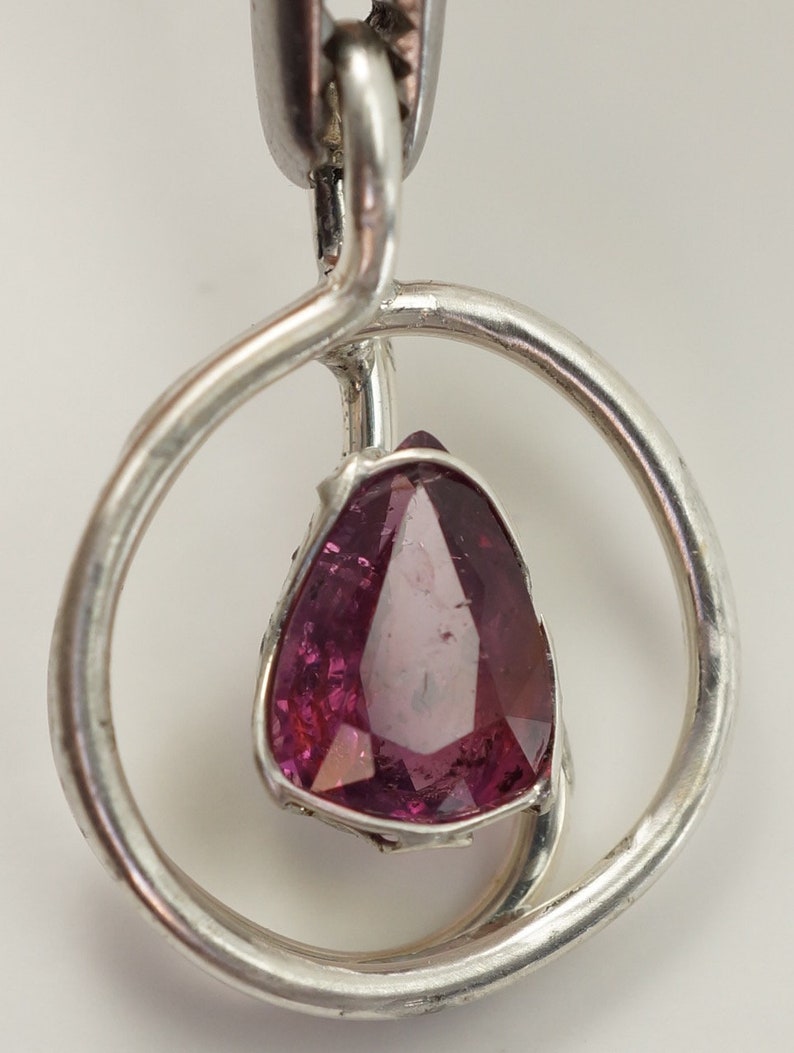 May include: A silver pendant featuring a pink gemstone set within a circular frame. The pendant is crafted with a twisted silver band.