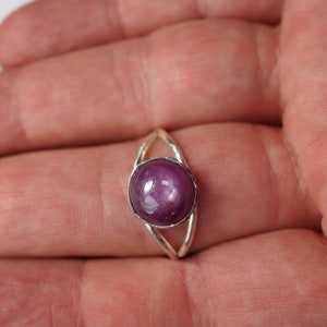 May include: A silver ring with a round, purple gemstone set in the center. The ring has a delicate, open design.