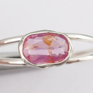 Puede incluir: Un anillo de plata con una piedra ovalada rosa engastada en un engaste de bisel. El anillo tiene una banda doble.