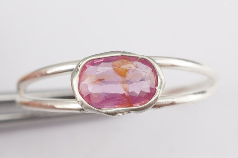 Puede incluir: Un anillo de plata con una piedra ovalada rosa engastada en un engaste de bisel. El anillo tiene una banda doble.