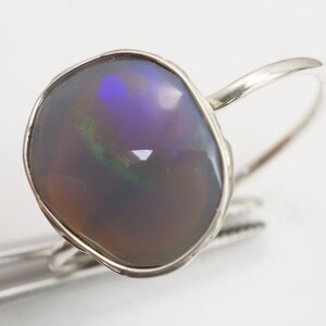 May include: A silver ring with a large, irregular shaped opal gemstone. The opal has a dark gray base with flashes of green, blue, and purple.