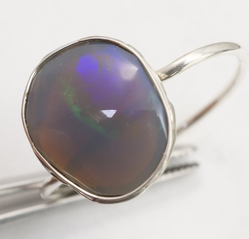 May include: A silver ring with a large, irregular shaped opal gemstone. The opal has a dark gray base with flashes of green, blue, and purple.