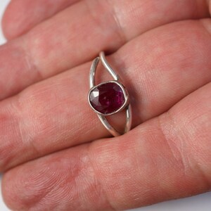 K&ouml;nnte beinhalten: Ein silberner Ring mit einem tief roten Edelstein, der in einem zarten, geschwungenen Band gefasst ist.