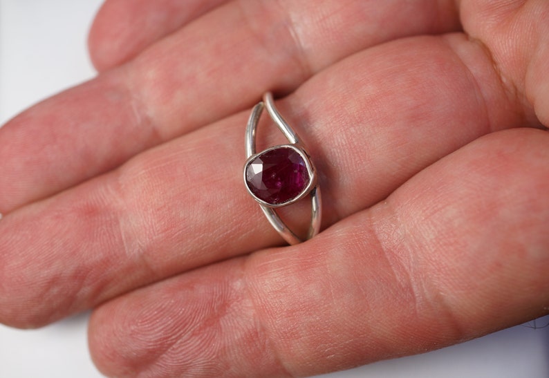 K&ouml;nnte beinhalten: Ein silberner Ring mit einem tief roten Edelstein, der in einem zarten, geschwungenen Band gefasst ist.