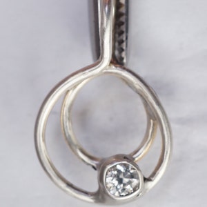 May include: A close-up of a silver pendant with a clear, round gemstone. The pendant has a unique design with two interlocking circles. The gemstone is set in a bezel setting, and the metal has a polished finish.