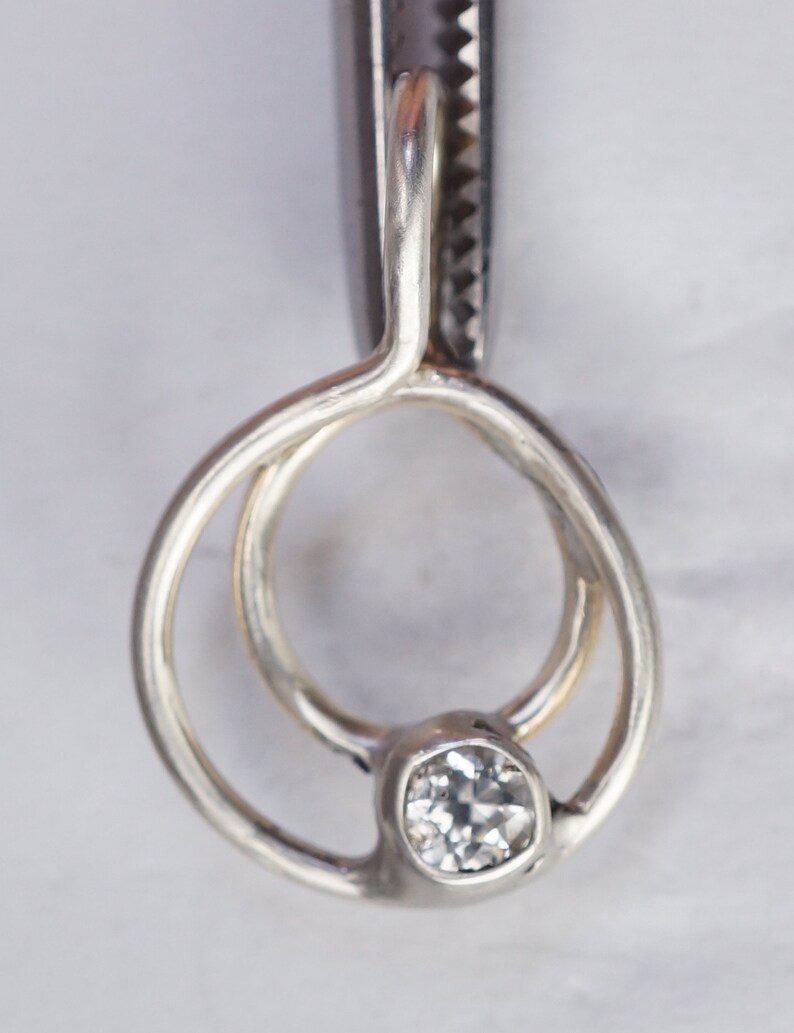 May include: A close-up of a silver pendant with a clear, round gemstone. The pendant has a unique design with two interlocking circles. The gemstone is set in a bezel setting, and the metal has a polished finish.