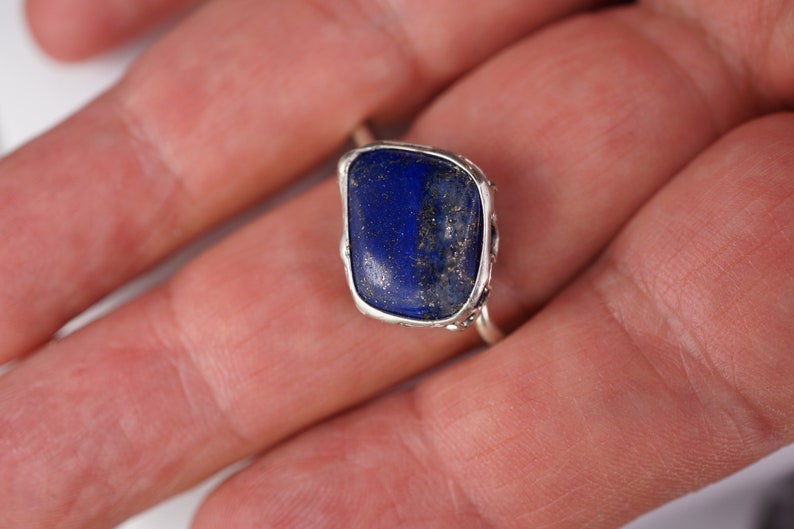 Puede incluir: Un anillo de plata con una gran piedra de lapisl&aacute;zuli azul oscuro engastada en un engaste de bisel. La piedra tiene una forma ligeramente irregular y est&aacute; rodeada por una delicada banda de plata.