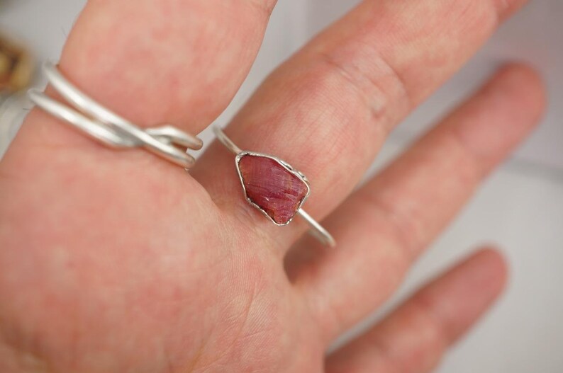 May include: A silver ring with a raw pink ruby gemstone set in the centre.