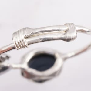 Puede incluir: Un anillo de plata con una piedra negra engastada en un engaste texturizado. El anillo está hecho de alambre que está retorcido y envuelto alrededor de la banda.
