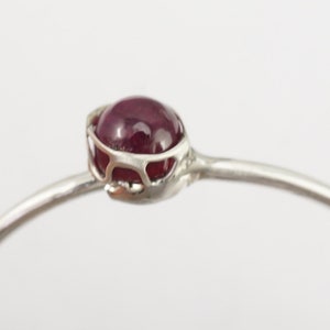 K&ouml;nnte beinhalten: Ein silbernes Armreif-Armband mit einem filigranen, durchbrochenen Design, das mit einem tief roten Edelstein verziert ist.