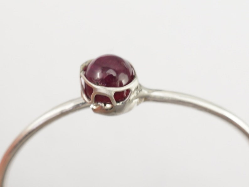 K&ouml;nnte beinhalten: Ein silbernes Armreif-Armband mit einem filigranen, durchbrochenen Design, das mit einem tief roten Edelstein verziert ist.