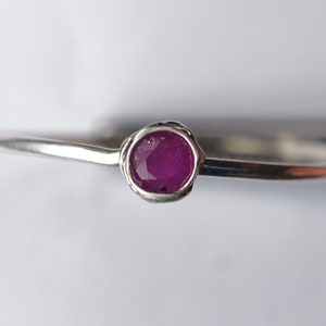 K&ouml;nnte beinhalten: Ein silberner Ring mit einem kleinen, runden, rosafarbenen Rubin-Edelstein, der in einer strukturierten Fassung sitzt.