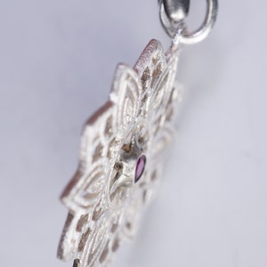 May include: A silver pendant featuring an intricate mandala design with a small, dark pink gemstone at its centre.