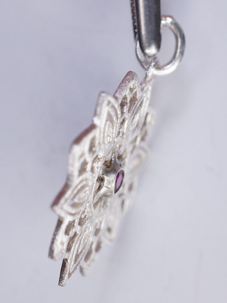 May include: A silver pendant featuring an intricate mandala design with a small, dark pink gemstone at its centre.