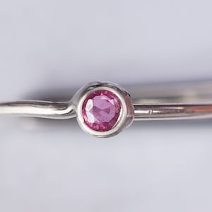 K&ouml;nnte beinhalten: Ein silberner Ring mit einem kleinen, runden, rosafarbenen Edelstein, der in einer strukturierten Fassung sitzt.