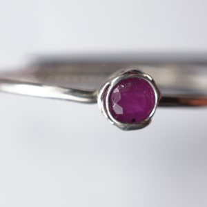 K&ouml;nnte beinhalten: Ein silberner Ring mit einem einzigen, runden, rosafarbenen Rubin-Edelstein, der in einem zarten, gedrehten Band gefasst ist.