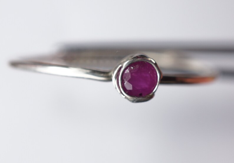 K&ouml;nnte beinhalten: Ein silberner Ring mit einem einzigen, runden, rosafarbenen Rubin-Edelstein, der in einem zarten, gedrehten Band gefasst ist.