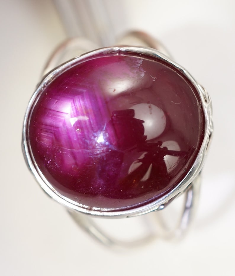 May include: A silver ring featuring a large, round, deep red ruby gemstone. The ruby has a star-like pattern in the centre.