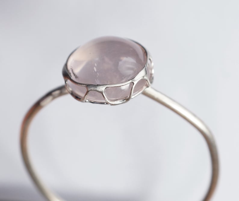 Puede incluir: Un anillo de plata con un delicado dise&ntilde;o de alambre que sostiene una piedra de cuarzo rosa p&aacute;lido.