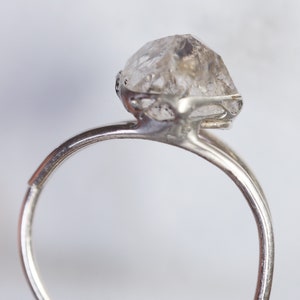May include: A silver ring with a clear, faceted, raw diamond gemstone set in a delicate, openwork design.