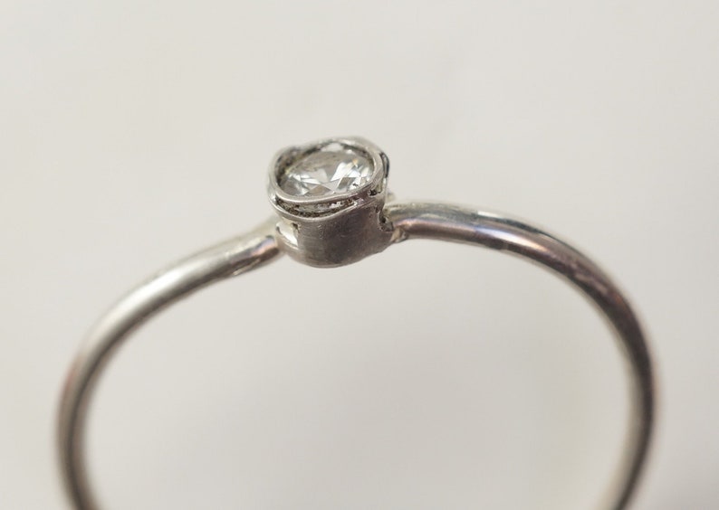 May include: A silver ring with a single round clear stone set in a delicate, textured band.
