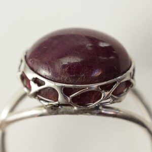 Pu&ograve; includere: Un anello d'argento con una grande pietra preziosa rotonda di colore rosso scuro incastonata in un design delicato e traforato.