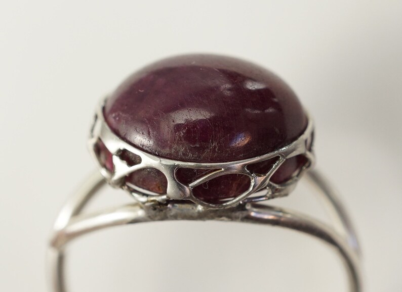 Pu&ograve; includere: Un anello d'argento con una grande pietra preziosa rotonda di colore rosso scuro incastonata in un design delicato e traforato.