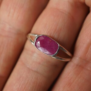 May include: A silver ring with a deep red ruby gemstone set in a bezel setting. The ring has a delicate, open design.