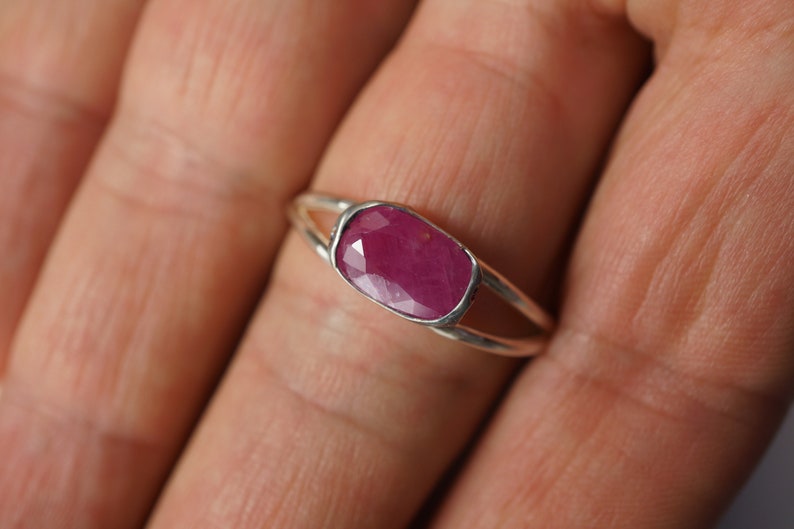 May include: A silver ring with a deep red ruby gemstone set in a bezel setting. The ring has a delicate, open design.