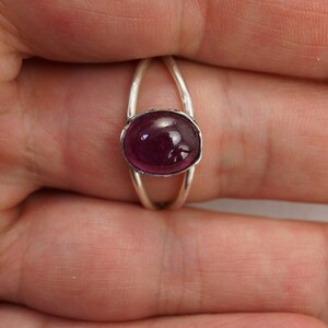 Puede incluir: Un anillo de plata con una gema grande, ovalada y de color magenta intenso. La banda del anillo tiene un dise&ntilde;o dividido, con dos arcos plateados delgados que se elevan para encontrarse con el engaste de la gema. La piedra pulida refleja la luz.