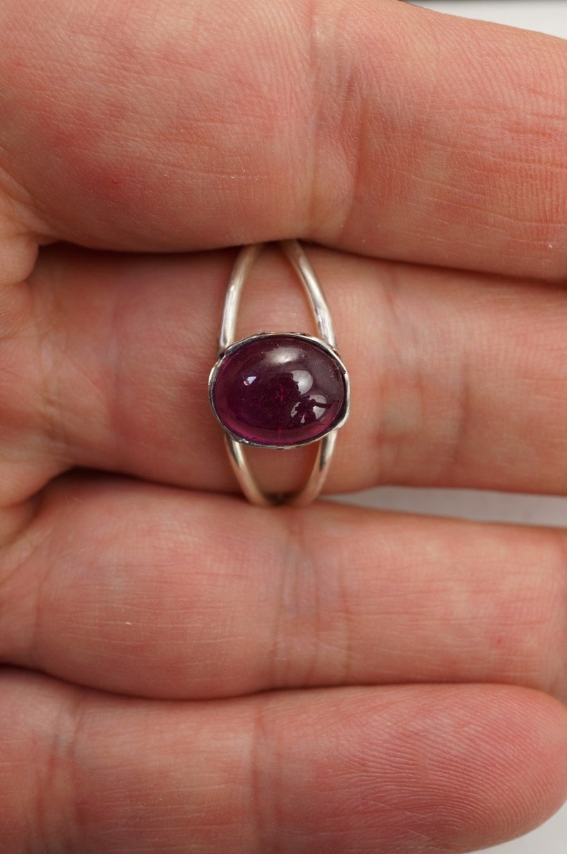 Puede incluir: Un anillo de plata con una gema grande, ovalada y de color magenta intenso. La banda del anillo tiene un dise&ntilde;o dividido, con dos arcos plateados delgados que se elevan para encontrarse con el engaste de la gema. La piedra pulida refleja la luz.