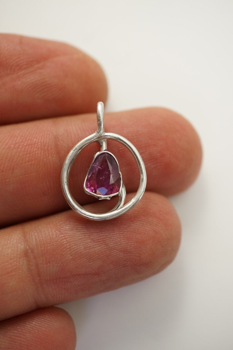 May include: A silver pendant featuring a pink gemstone set within a circular design.