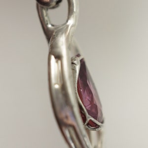 May include: A silver heart pendant featuring a purple gemstone set in the centre. The pendant has an intricate design and is attached to a silver chain.