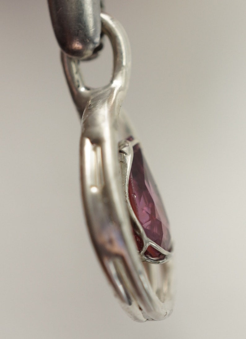 May include: A silver heart pendant featuring a purple gemstone set in the centre. The pendant has an intricate design and is attached to a silver chain.