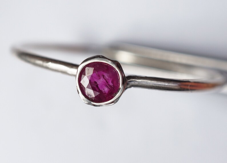 May include: A simple silver band ring with a small, round, faceted ruby gemstone.