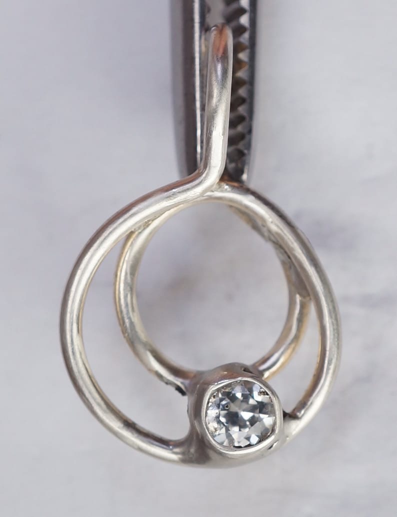 May include: A close-up of a silver pendant with a clear, round-cut gemstone. The pendant has a unique, open design with two interlocking loops. The gemstone is set in a bezel setting, and the metal has a polished finish.