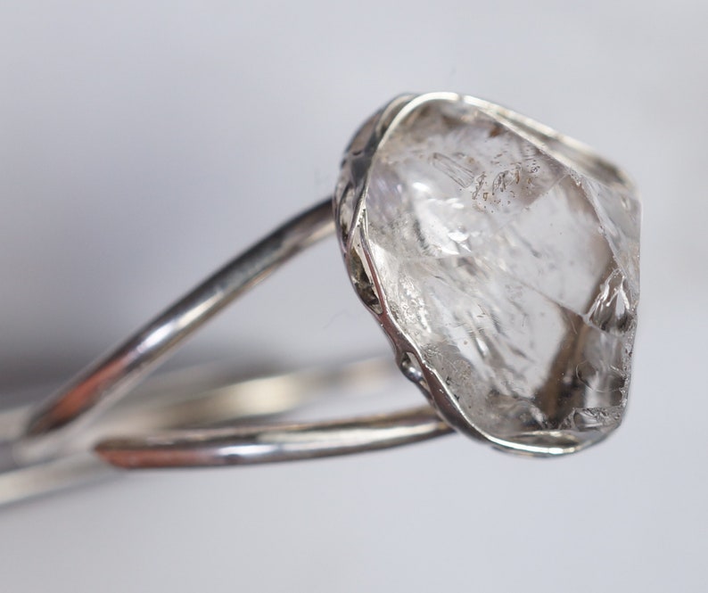 May include: A silver ring with a large, clear, faceted crystal set in the center. The crystal is rough and has a natural, unpolished look.