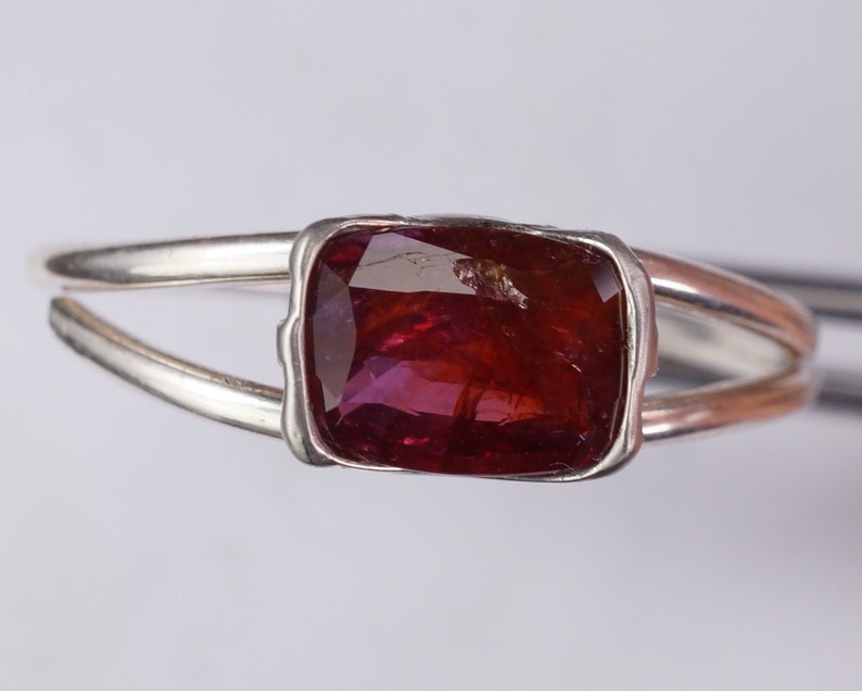 Pu&ograve; includere: Un anello d'argento con una pietra preziosa rossa incastonata in una montatura a castone. L'anello ha una gambo diviso.