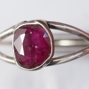 K&ouml;nnte beinhalten: Ein silberner Ring mit einem tief roten Rubin-Edelstein, der in einer Zargenfassung gefasst ist. Der Ring hat ein einzigartiges, organisches Design mit zwei B&auml;ndern, die den Stein umschlie&szlig;en.