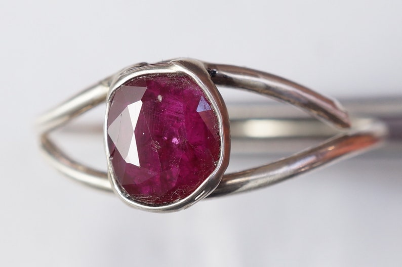 K&ouml;nnte beinhalten: Ein silberner Ring mit einem tief roten Rubin-Edelstein, der in einer Zargenfassung gefasst ist. Der Ring hat ein einzigartiges, organisches Design mit zwei B&auml;ndern, die den Stein umschlie&szlig;en.