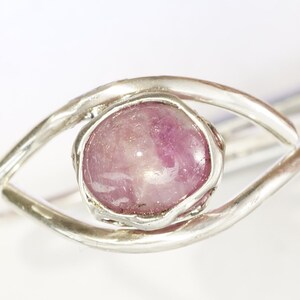May include: A silver ring featuring a pink gemstone set in a bezel setting. The ring is designed in an eye shape.
