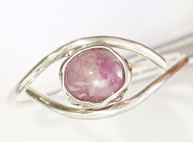 May include: A silver ring featuring a pink gemstone set in a bezel setting. The ring is designed in an eye shape.