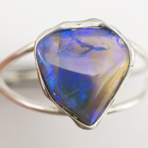 May include: A silver ring with a large, irregular shaped opal gemstone. The opal is a mix of blue, purple, and gold tones.